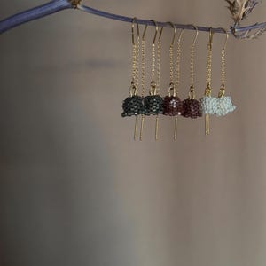 May include: Five pairs of beaded dangle earrings hanging from a branch. The earrings are made of small beads in shades of gray, burgundy, and white. The earrings are attached to gold chains.