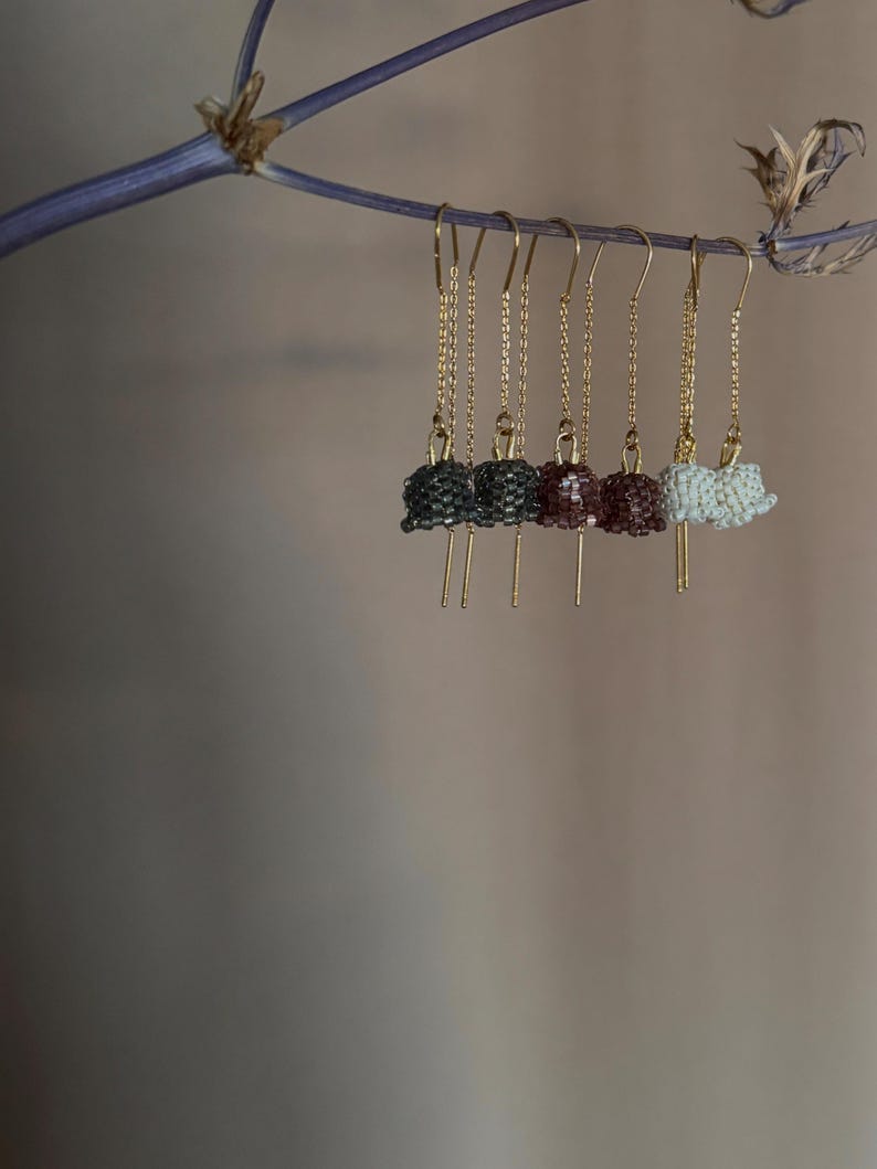 May include: Five pairs of beaded dangle earrings hanging from a branch. The earrings are made of small beads in shades of gray, burgundy, and white. The earrings are attached to gold chains.