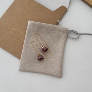 May include: A pair of gold earrings with small, beaded, bell-shaped charms in a light pink color. The earrings are displayed on a beige suede pouch with the text "LILIBEADS" printed on the pouch.