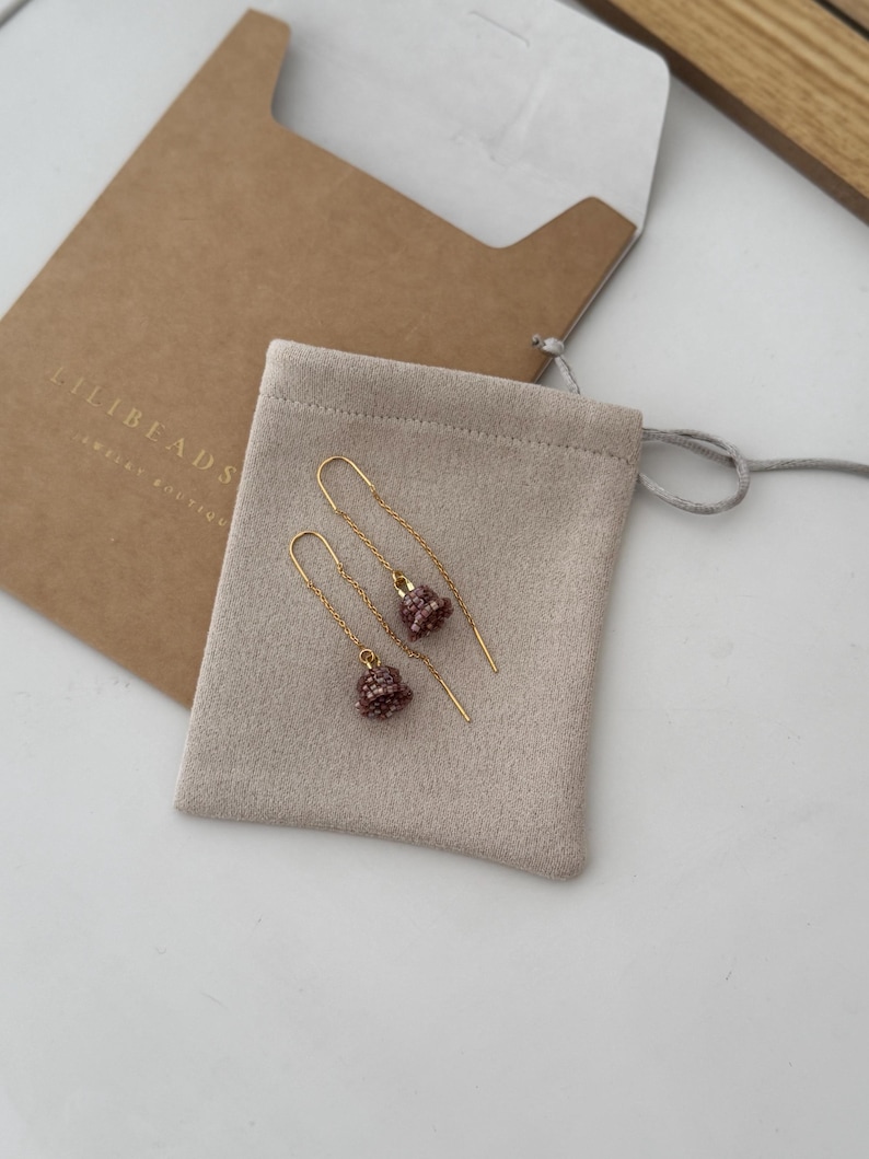 May include: A pair of gold earrings with small, beaded, bell-shaped charms in a light pink color. The earrings are displayed on a beige suede pouch with the text "LILIBEADS" printed on the pouch.