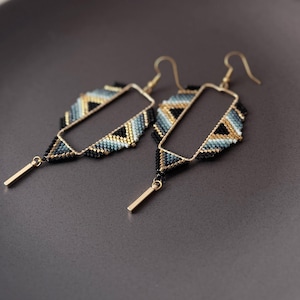 May include: A pair of beaded earrings with a geometric design. The earrings have a gold-coloured rectangular frame with a beaded feather-like design in black, blue, and gold. A small gold bar hangs from the bottom of each earring. The earrings are displayed on a dark grey plate.