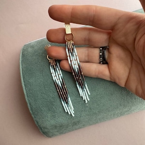 May include: A pair of long, beaded earrings with a gold hoop and a gold bar at the top. The beads are white, brown, and gray.