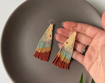 May include: A pair of long, beaded earrings with a gradient of colors, including beige, brown, light blue, and green. The earrings are made with small, round beads and have a tassel-like design.