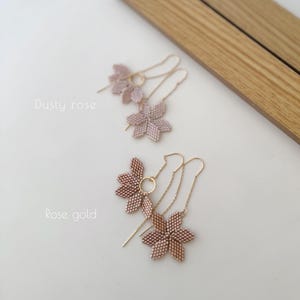May include: Two pairs of delicate earrings featuring a flower design. The earrings are available in two colors: dusty rose and rose gold. The earrings are made with small beads and have a dangling chain.