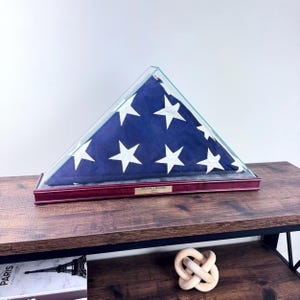 May include: A folded American flag in a triangular glass display case with a burgundy wooden base. The flag is blue with white stars and stripes. A small plaque reads "WILLIAM J. BROWN". The case sits on a dark wooden surface.