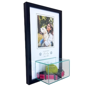 May include: A black rectangular wall-mounted shadow box frame featuring a photograph of a person with a dog. Below the photo, the text reads In Loving Memory Bailey 2012-2025 with paw prints. A glass display case holds a tennis ball and a red collar.