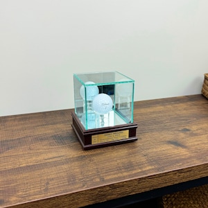 May include: A clear glass display case with a mirrored base, showcasing two Titleist golf balls. The case sits on a dark wooden base with a gold plaque. The plaque reads "JIMMY MOORE JUNE 2, 2024 NORTHERN HILLS GOLF COURSE HOLE IN ONE - 148 YDS - 7 IRON."