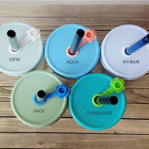 May include: Five colorful mason jar lids with straws. The lids are in shades of blue, green, and turquoise, each labeled with a different color name: Dew, Aqua, Icy Blue, Sage, and Turquoise. Each lid has a matching flower-shaped straw holder.