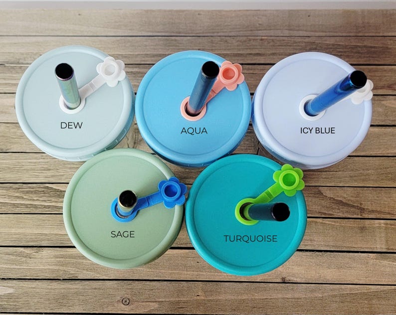 May include: Five colorful mason jar lids with straws. The lids are in shades of blue, green, and turquoise, each labeled with a different color name: Dew, Aqua, Icy Blue, Sage, and Turquoise. Each lid has a matching flower-shaped straw holder.