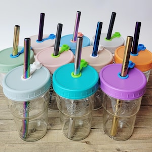 May include: A collection of clear glass mason jars with colorful lids and stainless steel straws. The lids are in various colors, including blue, pink, green, and purple. The straws have a metallic finish. The jars are arranged on a light-colored surface.