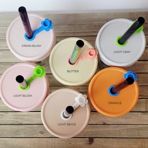 May include: Six mason jar lids in various colors, each with a straw and a flower-shaped holder. Colors include cream blush, light gray, light blush, butter, light beige, and orange. Each lid has a different colored straw and holder, with the color names printed on the lid.