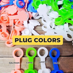 K&ouml;nnte beinhalten: Eine Sammlung von Silikonstopfen in den Farben Lachs, Limette, Wei&szlig; und Blau. Jeder Stopfen besteht aus einem kreisf&ouml;rmigen Ring, der an einer blumenf&ouml;rmigen Kappe befestigt ist. Der Text PLUG COLORS steht &uuml;ber vier einzelnen, beschrifteten Stopfen.
