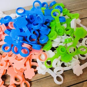 K&ouml;nnte beinhalten: Eine Sammlung von Kunststoff-Flaschenverschl&uuml;ssen in den Farben Blau, Gr&uuml;n, Wei&szlig; und Pfirsich. Jeder Verschluss besteht aus einem Ring, der &uuml;ber ein flexibles Band mit einem Stopfen verbunden ist, und einer blumenf&ouml;rmigen Lasche am Ende.
