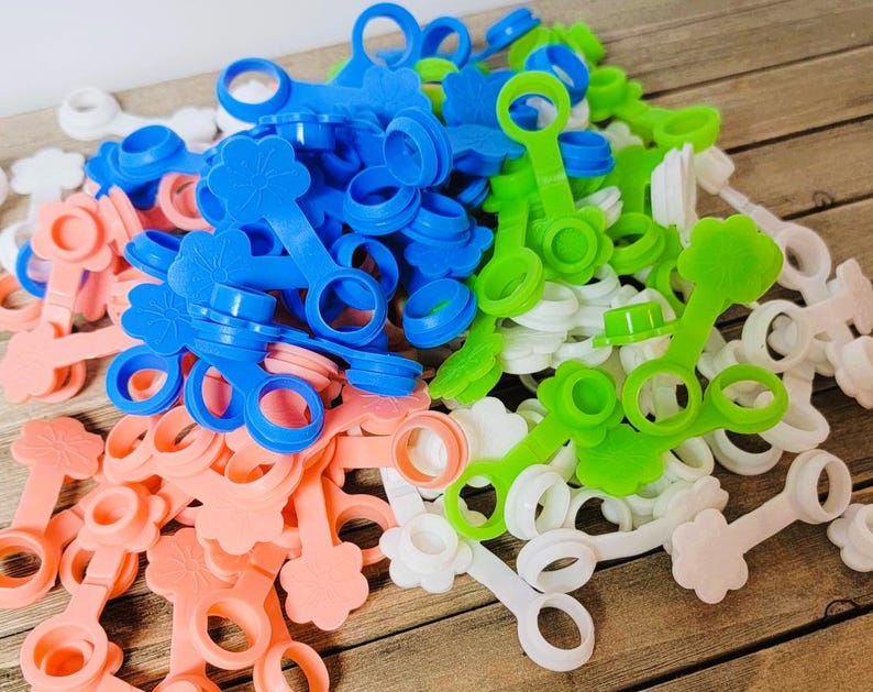 K&ouml;nnte beinhalten: Eine Sammlung von Kunststoff-Flaschenverschl&uuml;ssen in den Farben Blau, Gr&uuml;n, Wei&szlig; und Pfirsich. Jeder Verschluss besteht aus einem Ring, der &uuml;ber ein flexibles Band mit einem Stopfen verbunden ist, und einer blumenf&ouml;rmigen Lasche am Ende.
