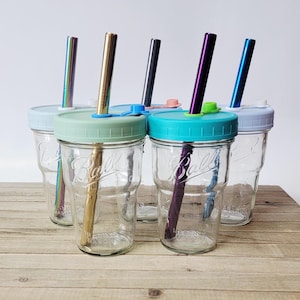 May include: Six clear glass mason jar mugs with colorful lids and metal straws. The lids are in shades of blue, green, and pink. The straws are in gold, silver, and iridescent colors. The jars are sitting on a wooden surface.