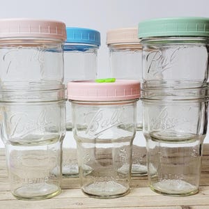 May include: A collection of clear glass Ball brand nesting mason jars with pastel-colored lids. The lids are pink, blue, peach, and green. The jars are stacked and arranged on a light-colored surface.
