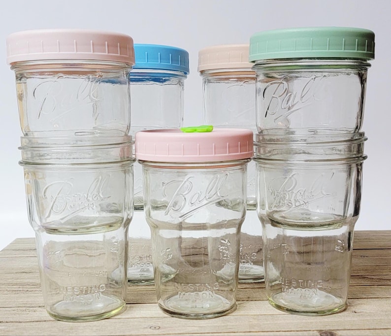 May include: A collection of clear glass Ball brand nesting mason jars with pastel-colored lids. The lids are pink, blue, peach, and green. The jars are stacked and arranged on a light-colored surface.