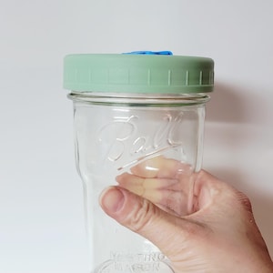 May include: A clear glass Ball mason jar with a light green lid and a blue insert. The jar is held by a hand and has the words "Nesting Mason" embossed on the bottom. The jar is empty.