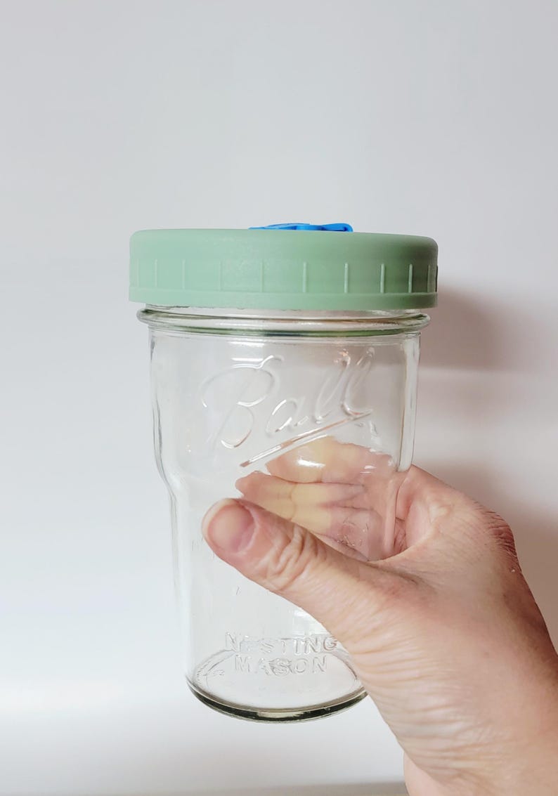 May include: A clear glass Ball mason jar with a light green lid and a blue insert. The jar is held by a hand and has the words "Nesting Mason" embossed on the bottom. The jar is empty.
