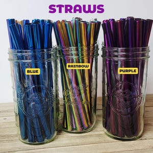 Pode incluir: Tr&ecirc;s frascos de vidro cheios de palhinhas reutiliz&aacute;veis azuis, arco-&iacute;ris e roxas. Os frascos t&ecirc;m r&oacute;tulos que dizem "BLUE", "RAINBOW" e "PURPLE". A palavra "STRAWS" est&aacute; escrita em roxo na parte superior da imagem.