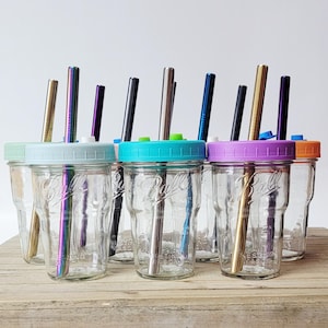 May include: A collection of clear glass mason jars with colorful lids and metal straws. The lids are in various colors, including blue, green, purple, and orange. The straws are in various colors, including gold, silver, and iridescent.