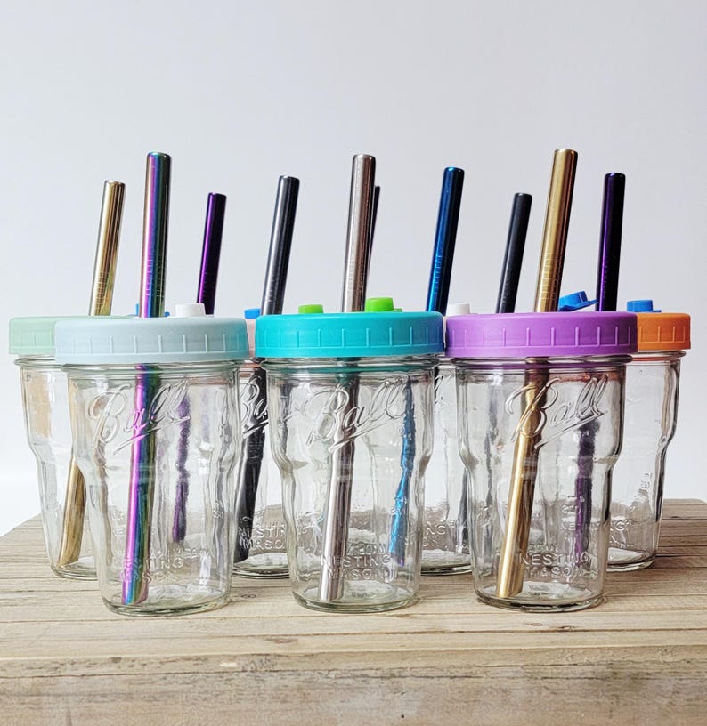 May include: A collection of clear glass mason jars with colorful lids and metal straws. The lids are in various colors, including blue, green, purple, and orange. The straws are in various colors, including gold, silver, and iridescent.