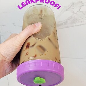 K&ouml;nnte beinhalten: Ein durchsichtiges Glas gef&uuml;llt mit Eiskaffee, mit einem violetten Deckel und einem kleinen gr&uuml;nen, kleeblattf&ouml;rmigen Silikonstopfen. Der Text auf dem Bild lautet: TURN ME UPSIDE DOWN I'M LEAKPROOF! und ein Pfeil zeigt auf SILICON PLUG.