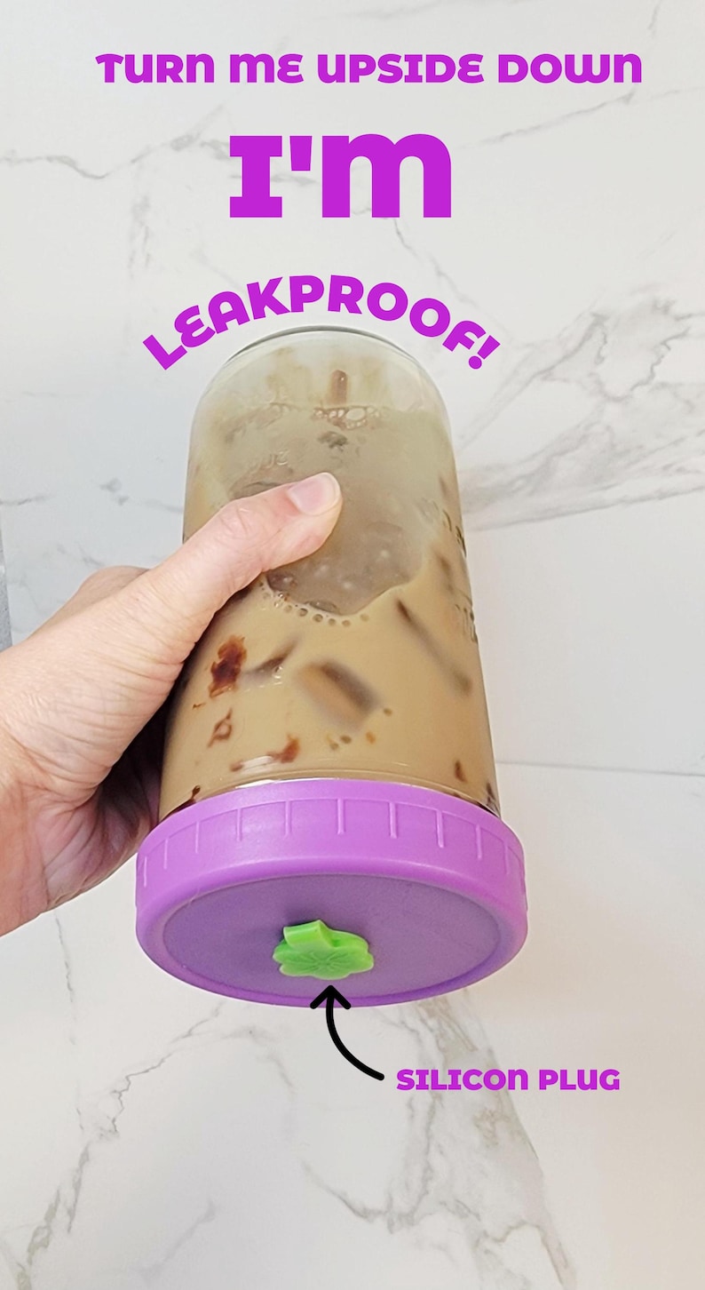 K&ouml;nnte beinhalten: Ein durchsichtiges Glas gef&uuml;llt mit Eiskaffee, mit einem violetten Deckel und einem kleinen gr&uuml;nen, kleeblattf&ouml;rmigen Silikonstopfen. Der Text auf dem Bild lautet: TURN ME UPSIDE DOWN I'M LEAKPROOF! und ein Pfeil zeigt auf SILICON PLUG.