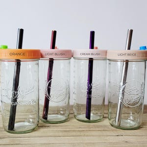 K&ouml;nnte beinhalten: Vier durchsichtige Einmachgl&auml;ser aus Glas mit gepr&auml;gtem Logo Alpine Canning Co. Jedes Glas hat einen farbigen Deckel mit der Aufschrift Orange, Light Blush, Cream Blush oder Light Beige sowie einen passenden Metallstrohhalm und Silikonstopfen.