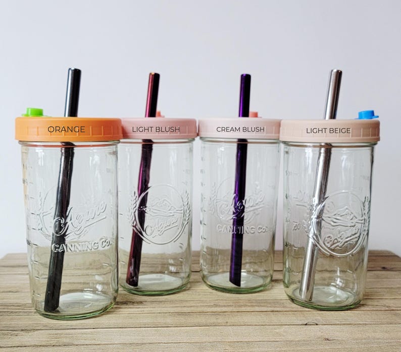 K&ouml;nnte beinhalten: Vier durchsichtige Einmachgl&auml;ser aus Glas mit gepr&auml;gtem Logo Alpine Canning Co. Jedes Glas hat einen farbigen Deckel mit der Aufschrift Orange, Light Blush, Cream Blush oder Light Beige sowie einen passenden Metallstrohhalm und Silikonstopfen.