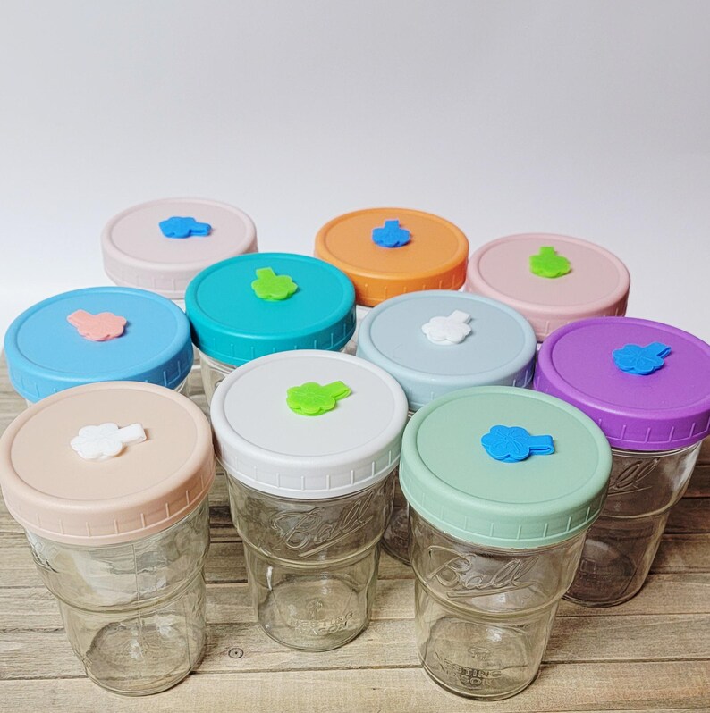 May include: A collection of clear glass Ball jars with colorful lids. The lids are pastel shades of pink, blue, green, orange, and purple, each topped with a small, decorative cloud-shaped accent. The jars are empty and ready for use.
