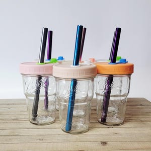 May include: Clear glass mason jars with pastel-colored lids and metal straws. The jars have pink, beige, and orange lids, with straws in various colors. The jars are sitting on a wooden surface.