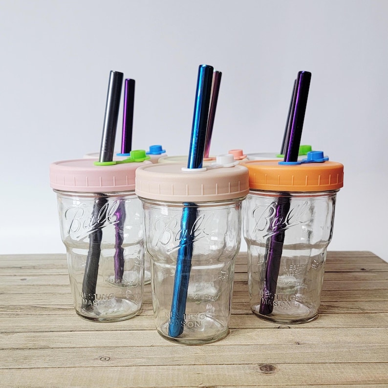May include: Clear glass mason jars with pastel-colored lids and metal straws. The jars have pink, beige, and orange lids, with straws in various colors. The jars are sitting on a wooden surface.