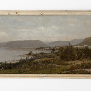 May include: A framed oil painting depicting a landscape scene with a lake, mountains, and trees. The painting is in a light wood frame and has a muted color palette.
