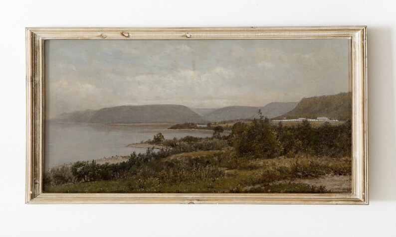 May include: A framed oil painting depicting a landscape scene with a lake, mountains, and trees. The painting is in a light wood frame and has a muted color palette.