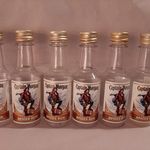 Puede incluir: Seis botellas en miniatura de ron especiado Captain Morgan Original. Cada botella transparente tiene una tapa dorada y una etiqueta con una ilustración de pirata. Las botellas están dispuestas en fila.