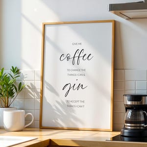 Op de afbeelding: Een ingelijste print met de tekst "Give me coffee to change the things I can & gin to accept the things I can't." De print heeft een houten frame en wordt in een keuken weergegeven. Een witte mok en een koffiezetapparaat zijn ook zichtbaar.