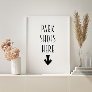 以下が含まれることがあります： 「PARK SHOES HERE」の黒い文字と下向きの黒い矢印が描かれた白い額縁のサイン。白いキャビネットの上に飾られており、装飾としてドライフラワーとパンパスグラスが花瓶に入っています。