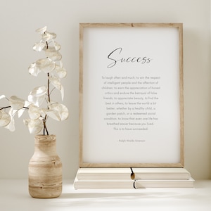 Puede incluir: Una impresi&oacute;n enmarcada con la palabra "&Eacute;xito" y una cita de Ralph Waldo Emerson. La impresi&oacute;n est&aacute; en un marco de madera clara, apoyada en una pila de libros. Un jarr&oacute;n decorativo con flores blancas est&aacute; a la izquierda.