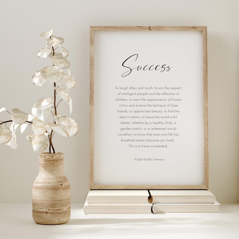 Puede incluir: Una impresi&oacute;n enmarcada con la palabra "&Eacute;xito" y una cita de Ralph Waldo Emerson. La impresi&oacute;n est&aacute; en un marco de madera clara, apoyada en una pila de libros. Un jarr&oacute;n decorativo con flores blancas est&aacute; a la izquierda.