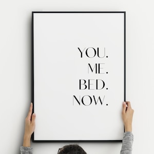 May include: A black framed print featuring the words "YOU. ME. BED. NOW." in a minimalist font. The print is held by a person against a white background. The artwork is a modern, romantic statement piece for a bedroom.