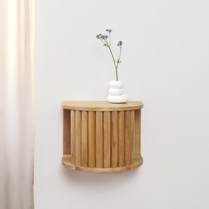 Könnte beinhalten: Ein wandmontiertes Holzregal mit einer gebogenen Vorderseite und einem Lamellendesign. Das Regal ist aus hellem Holz gefertigt und hat eine kleine Vase mit Blumen darauf.