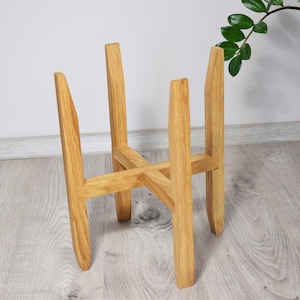 May include: A natural wood plant stand with a four-legged design. The stand is made of light-colored wood and has a simple, modern aesthetic. The stand is designed to hold a potted plant and is suitable for indoor use.