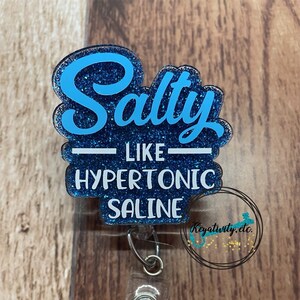 K&ouml;nnte beinhalten: Ein blau glitzernder Ausweis-Clip mit den Worten "Salty like Hypertonic Saline" in wei&szlig;er und blauer Schrift. Der Ausweis-Clip hat einen transparenten Kunststoffclip und einen dekorativen Schmuckstein.