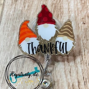 May include: A clear plastic badge holder with a gold glitter background and the word "Thankful" in gold glitter lettering. Three gnomes with red, orange, and brown hats are on the holder.