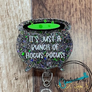 May include: A Halloween-themed badge reel featuring a cauldron design with the text "IT'S JUST A BUNCH OF HOCUS POCUS!". The cauldron is black with green accents and glitter. The badge reel has a clear plastic strap and a metal clip.