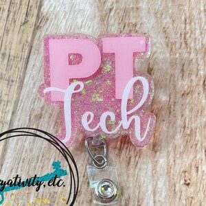 May include: A pink glitter badge reel featuring the text PT Tech in bold block letters and white script. The badge reel has a clear plastic strap with a metal snap and a small rhinestone accent at the top of the clip mechanism.