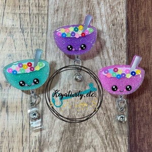 May include: Three glittery badge reels in teal, purple, and pink, each shaped like a bowl of cereal with a spoon. The bowls have smiling faces and are filled with colorful, round cereal pieces. The reels have a clear plastic strap and a metal clip.