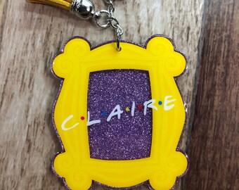 May include: A yellow acrylic keychain in the shape of a picture frame with the name "CLAIRE" and the word "FRIENDS" in the center. The frame has a purple glitter background and a yellow tassel.