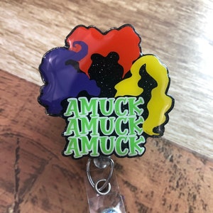 May include: A badge reel with a design featuring three colorful silhouettes of hair, with the text "AMUCK" in green. The reel has a clear plastic strap and a decorative, glittery button.